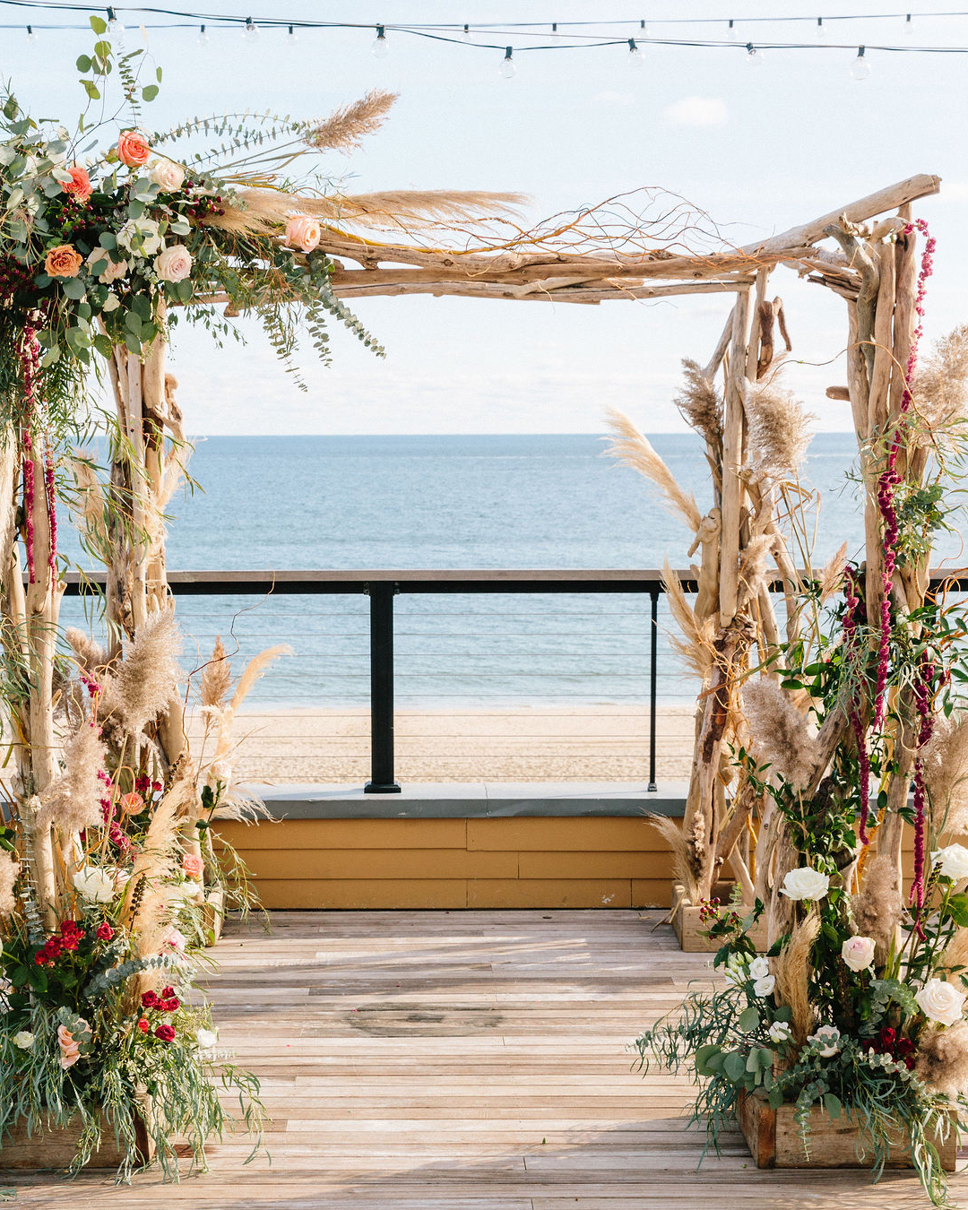 Beachfront wedding arch adorned with flowers and pampas grass, wooden deck and ocean in the background.